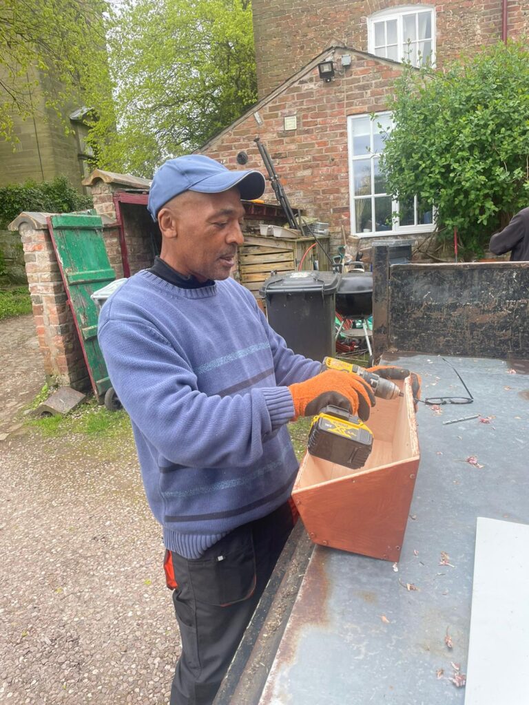 Photograph of a man in a cap drilling holes in a swift box ready for installation on the wall of the brick house behind him. Swift conservation in action!