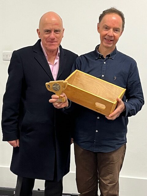 Two white men stand side by side. One is holding a small wooden trophy and a wooden swift box.