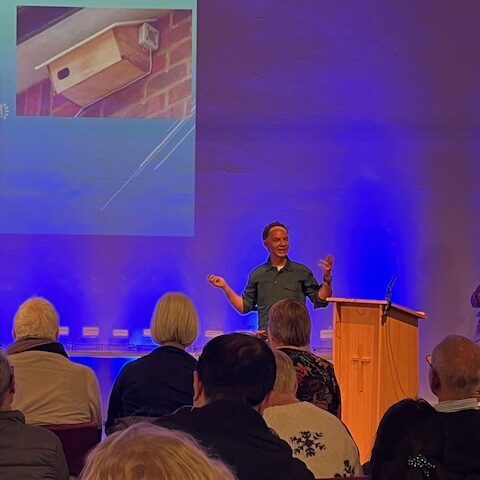 SMS Secretary Andy stands in front of a seated audience making a speech with a project image of a swift box on the wall behind him.